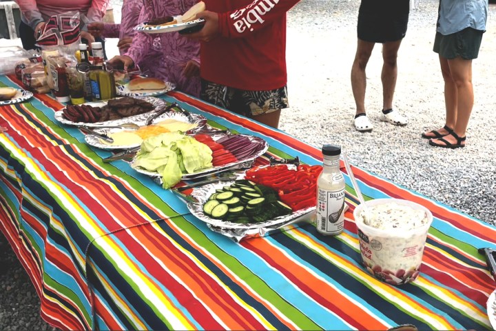 Outdoor picnic table with colorful cloth, food trays, and people serving themselves.