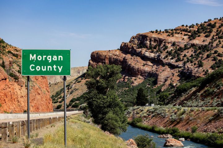 Morgan County sign beside road with red rock cliffs and greenery.
