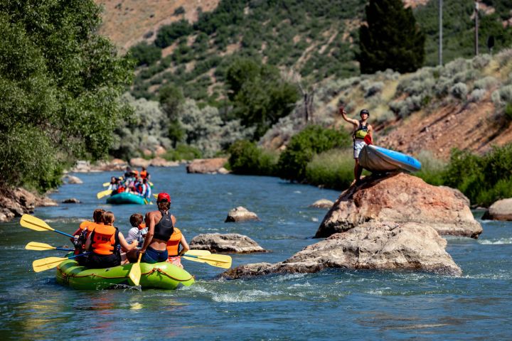 People in rafts paddling down a river, one person standing on a rock holding a kayak.