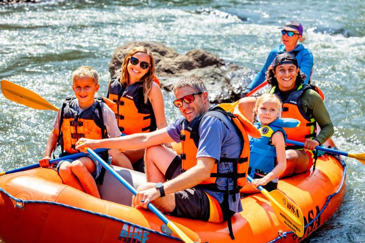 Group of people wearing life vests rafting on a river in an orange raft, smiling at the camera.