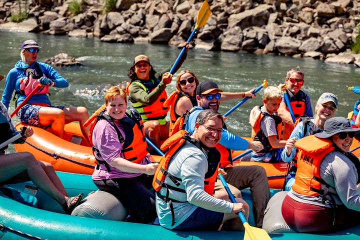 Group smiling and paddling in an inflatable raft on a river, wearing life jackets and helmets.
