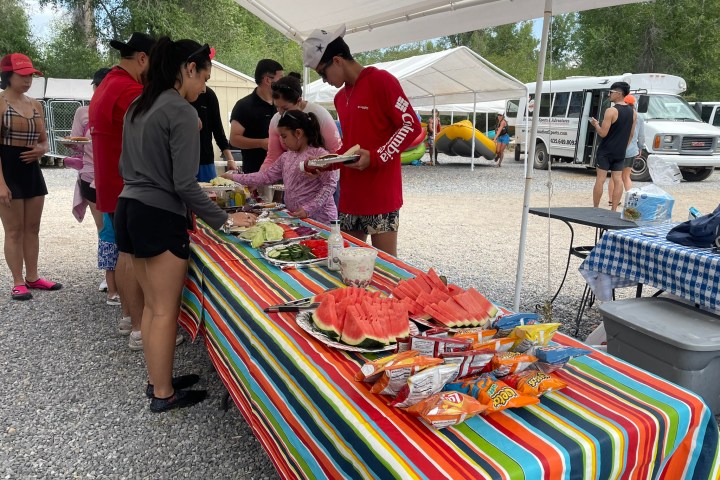 People serving food from a colorful table under a canopy, with chips and watermelon visible.