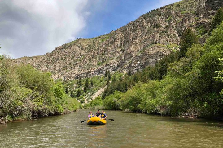 Yellow raft with people on the river, surrounded by lush green trees and rocky cliffs.