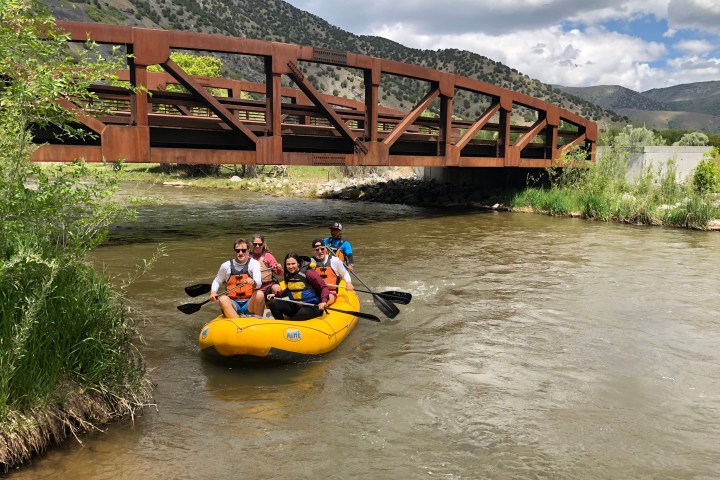 Group rafting under a wooden bridge on a sunny day with mountains in the background.