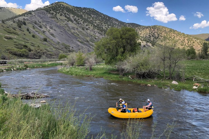 Three people rafting on a river with hills and trees in the background.