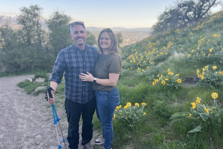 Couple smiles on a hillside path with yellow flowers and trees in the background.