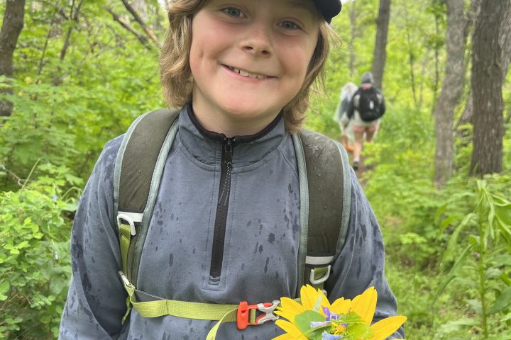 Child holding a yellow flower, wearing a cap and backpack in a forest.