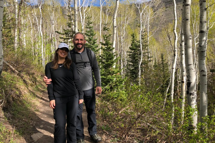 Two people smiling on a forest trail with tall trees and mountains in the background.