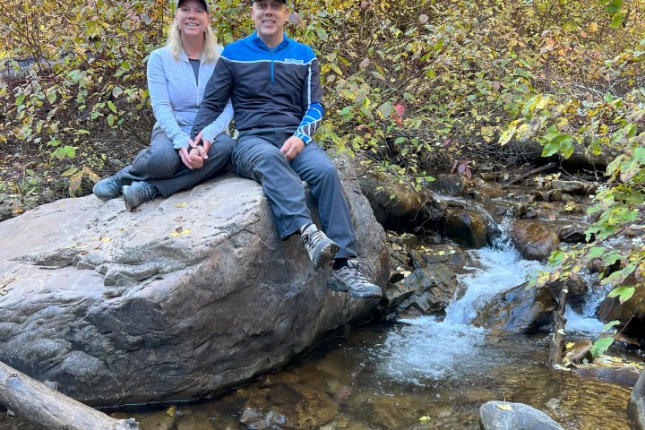 Couple sitting on rock by stream in autumn woods, holding hands.